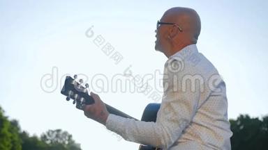 一个秃顶的人在公园里弹吉他唱歌。 吉他手触摸吉他弦。 中弹。